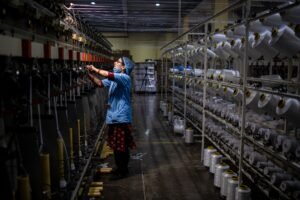 Masked female worker operating textile machinery in a factory setting. Indoor shot.